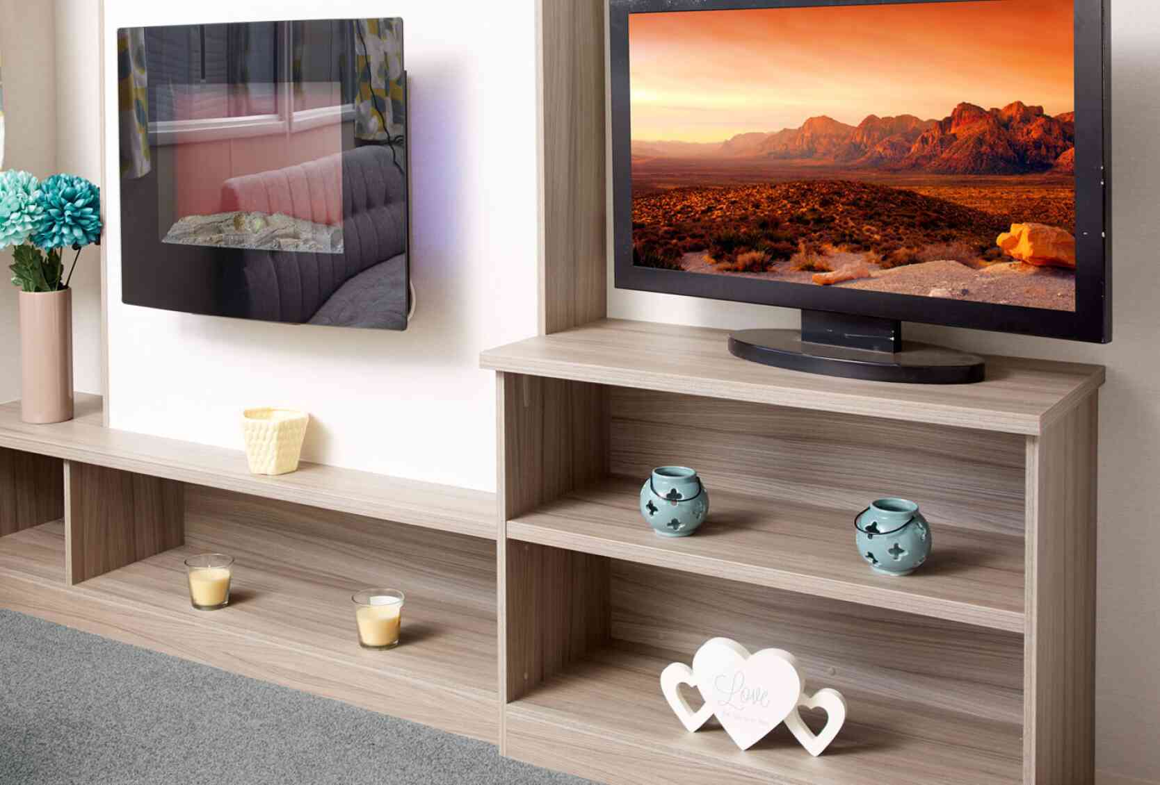 The image shows a close-up of a modern caravan or mobile home entertainment area. On the left, there is a sleek wall-mounted electric fireplace with a reflective glass surface, showing the reflection of a sofa and patterned curtains. Below it is a long wooden shelf holding a vase with blue artificial flowers, a small yellow candle holder, and two tealight candles. On the right, a flat-screen TV displays a vivid landscape of rocky mountains under an orange sky. The TV sits on a light wood shelving unit that contains two turquoise decorative candle holders on the middle shelf and a white heart-shaped ornament with the words “Love the life you live” on the bottom shelf. The floor is carpeted in soft grey.