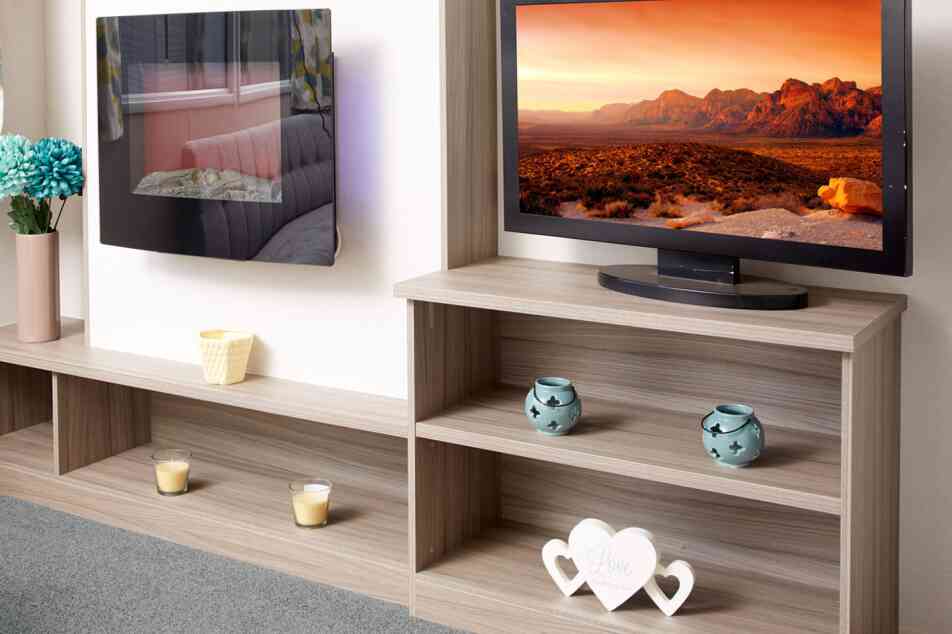 The image shows a close-up of a modern caravan or mobile home entertainment area. On the left, there is a sleek wall-mounted electric fireplace with a reflective glass surface, showing the reflection of a sofa and patterned curtains. Below it is a long wooden shelf holding a vase with blue artificial flowers, a small yellow candle holder, and two tealight candles. On the right, a flat-screen TV displays a vivid landscape of rocky mountains under an orange sky. The TV sits on a light wood shelving unit that contains two turquoise decorative candle holders on the middle shelf and a white heart-shaped ornament with the words “Love the life you live” on the bottom shelf. The floor is carpeted in soft grey.