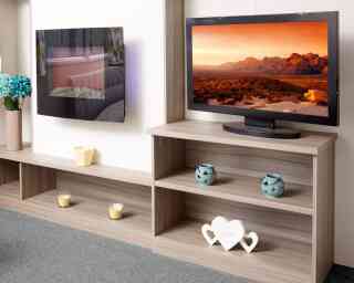The image shows a close-up of a modern caravan or mobile home entertainment area. On the left, there is a sleek wall-mounted electric fireplace with a reflective glass surface, showing the reflection of a sofa and patterned curtains. Below it is a long wooden shelf holding a vase with blue artificial flowers, a small yellow candle holder, and two tealight candles. On the right, a flat-screen TV displays a vivid landscape of rocky mountains under an orange sky. The TV sits on a light wood shelving unit that contains two turquoise decorative candle holders on the middle shelf and a white heart-shaped ornament with the words “Love the life you live” on the bottom shelf. The floor is carpeted in soft grey.