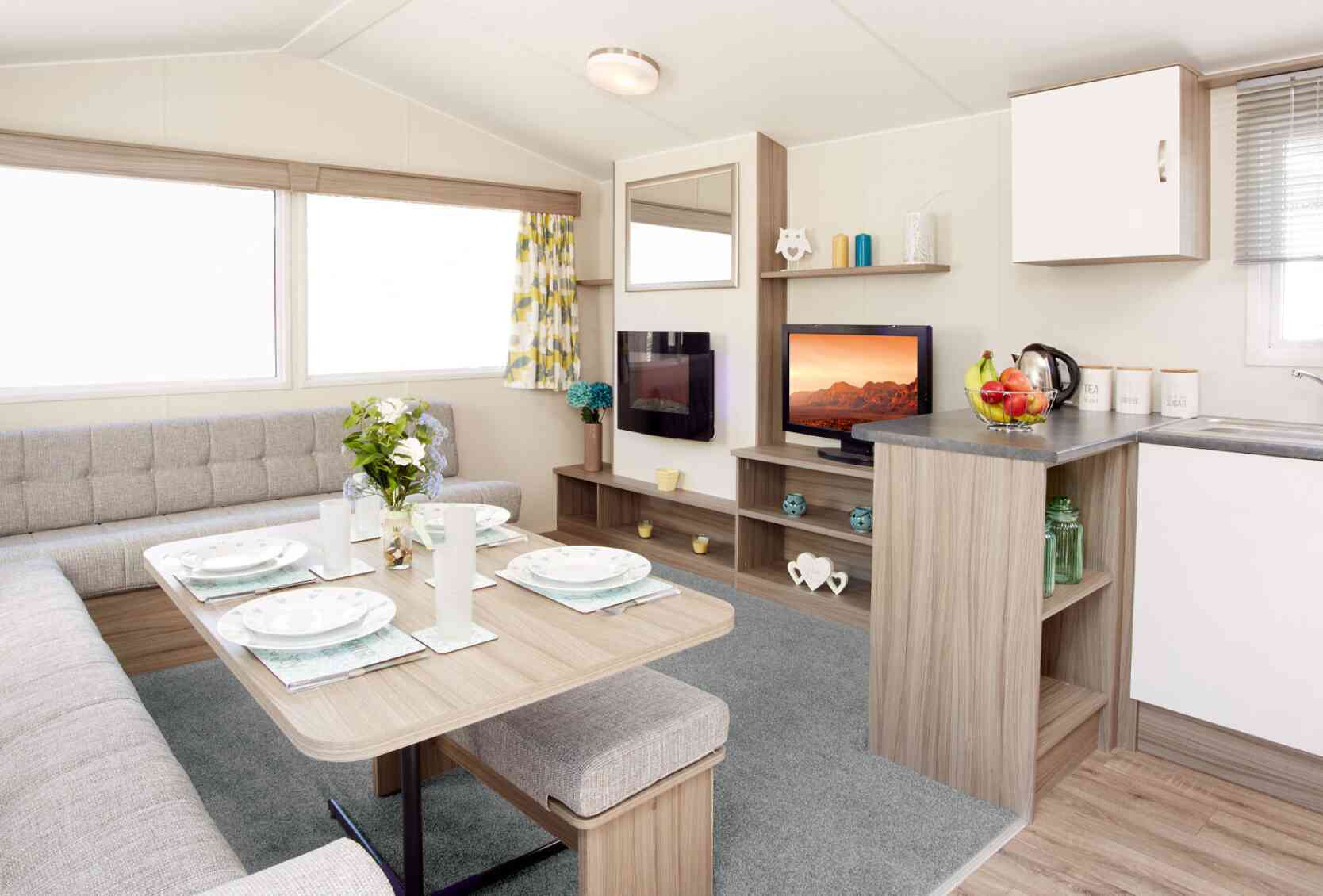 The image shows the interior of a modern caravan or mobile home. It features an L-shaped light-grey upholstered seating area surrounding a wooden dining table set with four plates, glasses, and placemats. A vase of fresh flowers sits in the centre of the table. To the right, there’s a compact kitchen area with a grey countertop holding a fruit bowl, a kettle, and labelled storage canisters. Behind the counter is a small entertainment area with a flat-screen TV displaying a scenic landscape, shelves with decorative items, and a large window with patterned yellow-and-grey curtains letting in natural light. The floor is a mix of light wood and a soft grey carpet in the lounge area.