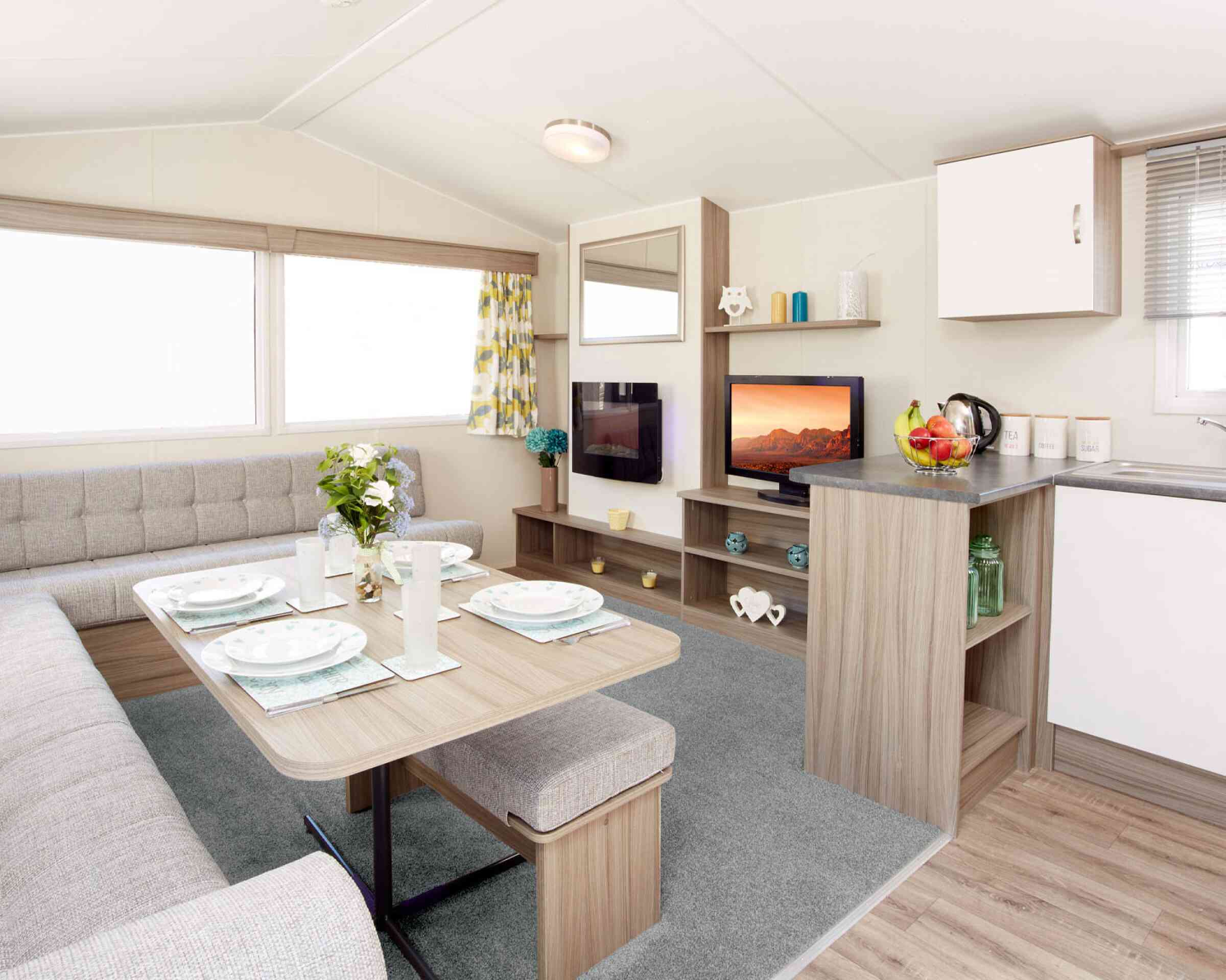 The image shows the interior of a modern caravan or mobile home. It features an L-shaped light-grey upholstered seating area surrounding a wooden dining table set with four plates, glasses, and placemats. A vase of fresh flowers sits in the centre of the table. To the right, there’s a compact kitchen area with a grey countertop holding a fruit bowl, a kettle, and labelled storage canisters. Behind the counter is a small entertainment area with a flat-screen TV displaying a scenic landscape, shelves with decorative items, and a large window with patterned yellow-and-grey curtains letting in natural light. The floor is a mix of light wood and a soft grey carpet in the lounge area.