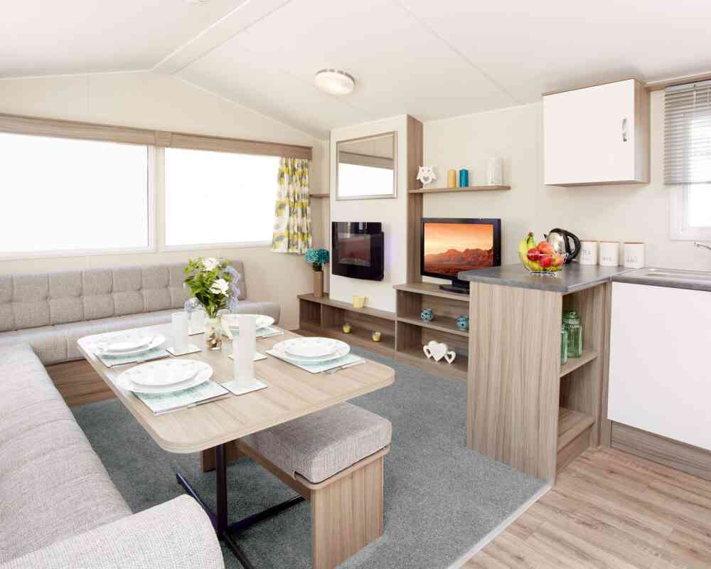 The image shows the interior of a modern caravan or mobile home. It features an L-shaped light-grey upholstered seating area surrounding a wooden dining table set with four plates, glasses, and placemats. A vase of fresh flowers sits in the centre of the table. To the right, there’s a compact kitchen area with a grey countertop holding a fruit bowl, a kettle, and labelled storage canisters. Behind the counter is a small entertainment area with a flat-screen TV displaying a scenic landscape, shelves with decorative items, and a large window with patterned yellow-and-grey curtains letting in natural light. The floor is a mix of light wood and a soft grey carpet in the lounge area.