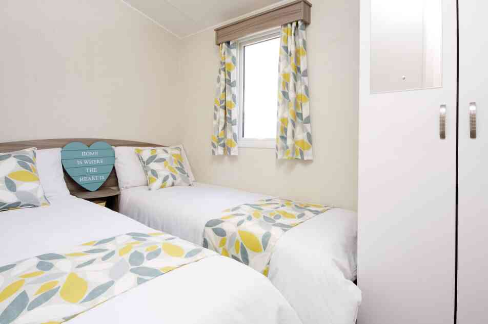 A cosy second bedroom featuring two beds, white walls, and yellow-green curtains.