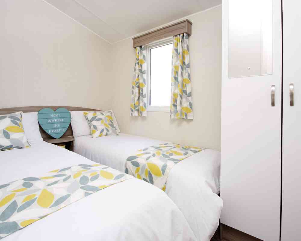 A cosy second bedroom featuring two beds, white walls, and yellow-green curtains.