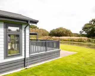 A modern cabin with a gray and light blue exterior, featuring a wooden deck overlooking a grassy area and a serene body of water in the background. Trees and lush greenery are visible in the distance under a partly cloudy sky.