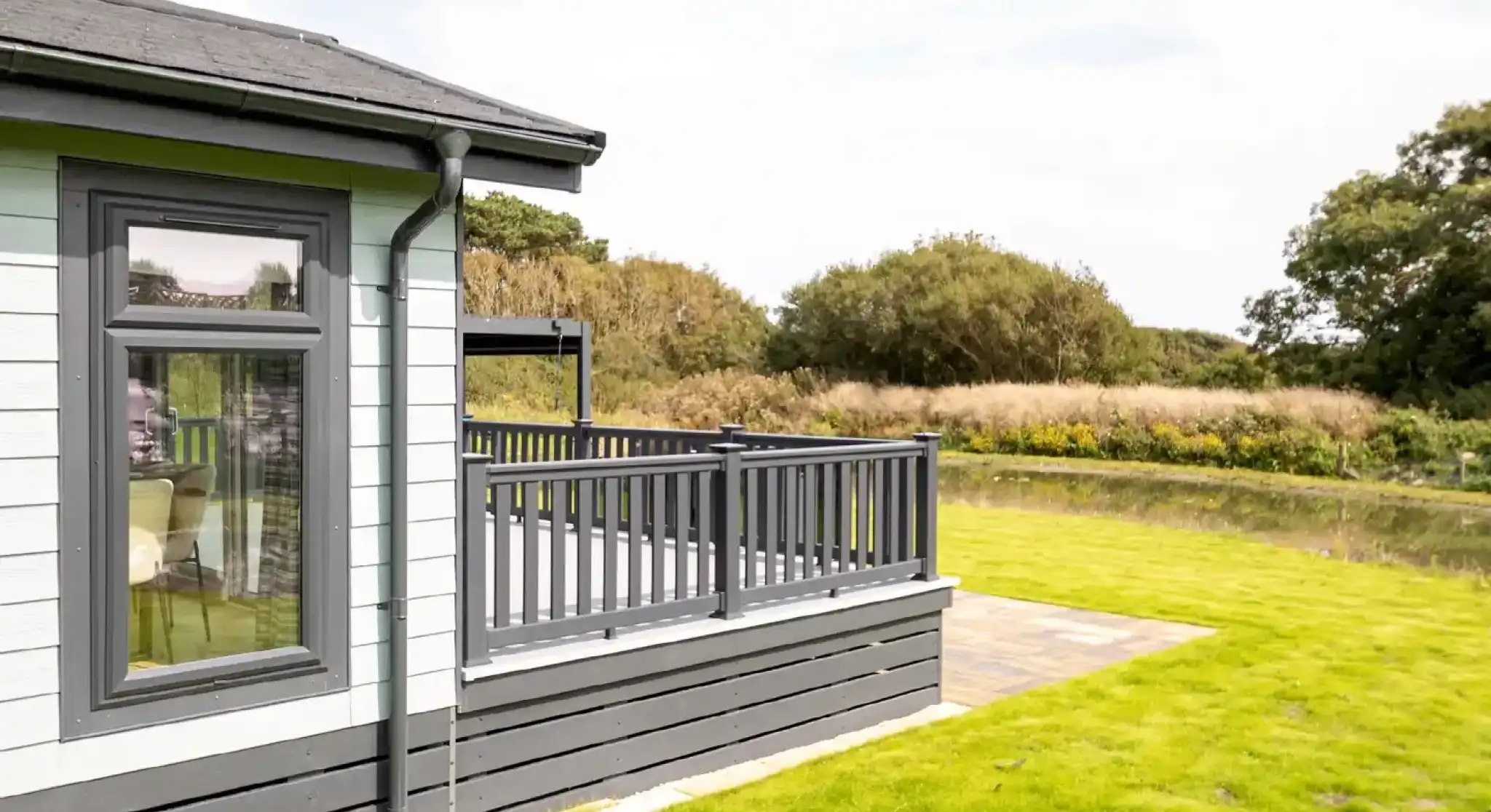 A modern cabin with a gray and light blue exterior, featuring a wooden deck overlooking a grassy area and a serene body of water in the background. Trees and lush greenery are visible in the distance under a partly cloudy sky.