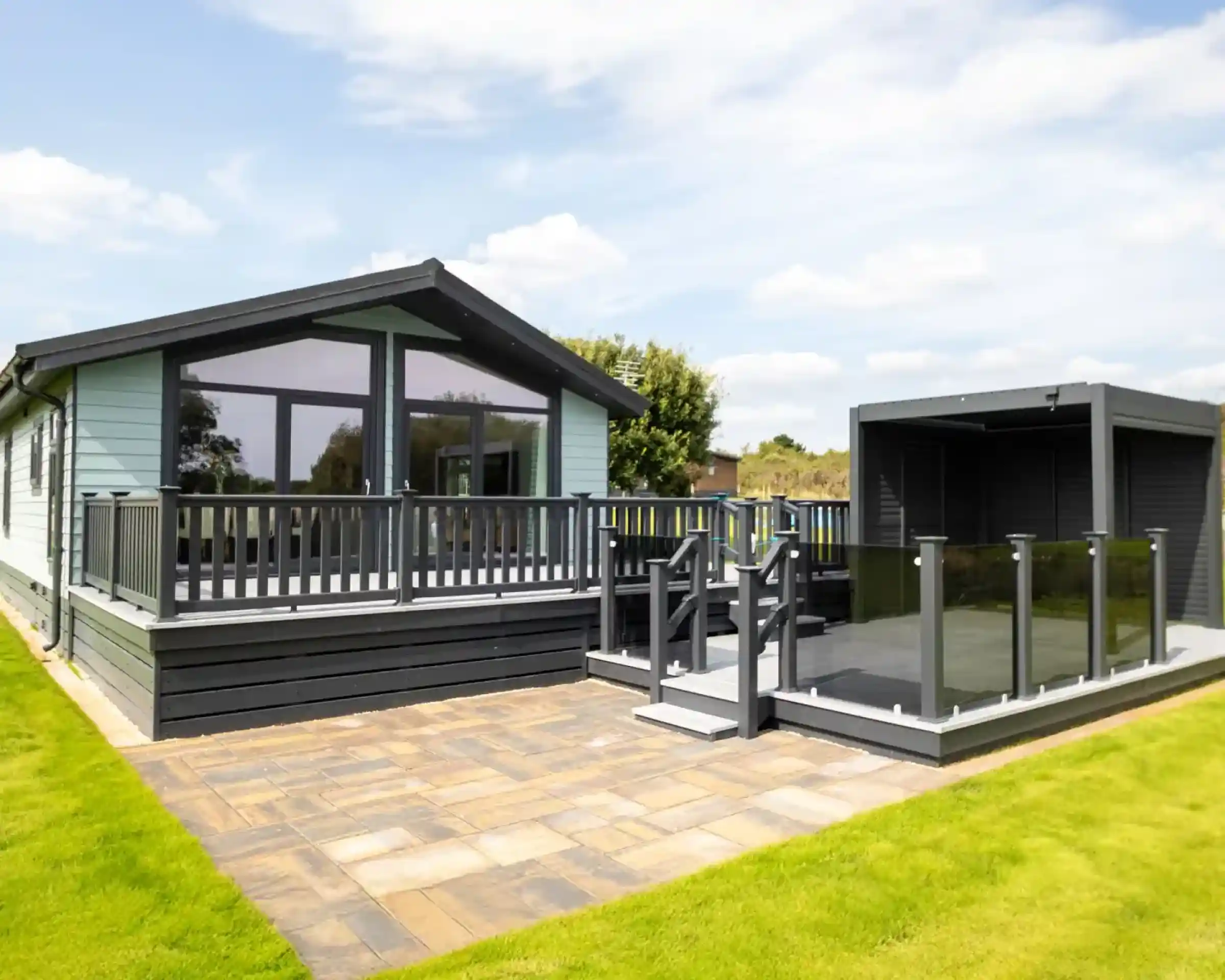 Modern light blue holiday home with a grey wraparound deck and glass balustrades, adjacent to a dark grey outdoor pergola.