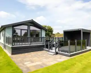 Modern light blue holiday home with a grey wraparound deck and glass balustrades, adjacent to a dark grey outdoor pergola.
