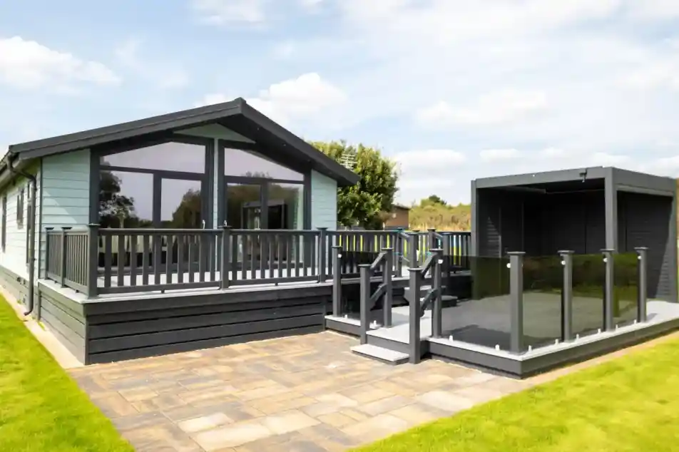 Modern light blue holiday home with a grey wraparound deck and glass balustrades, adjacent to a dark grey outdoor pergola.