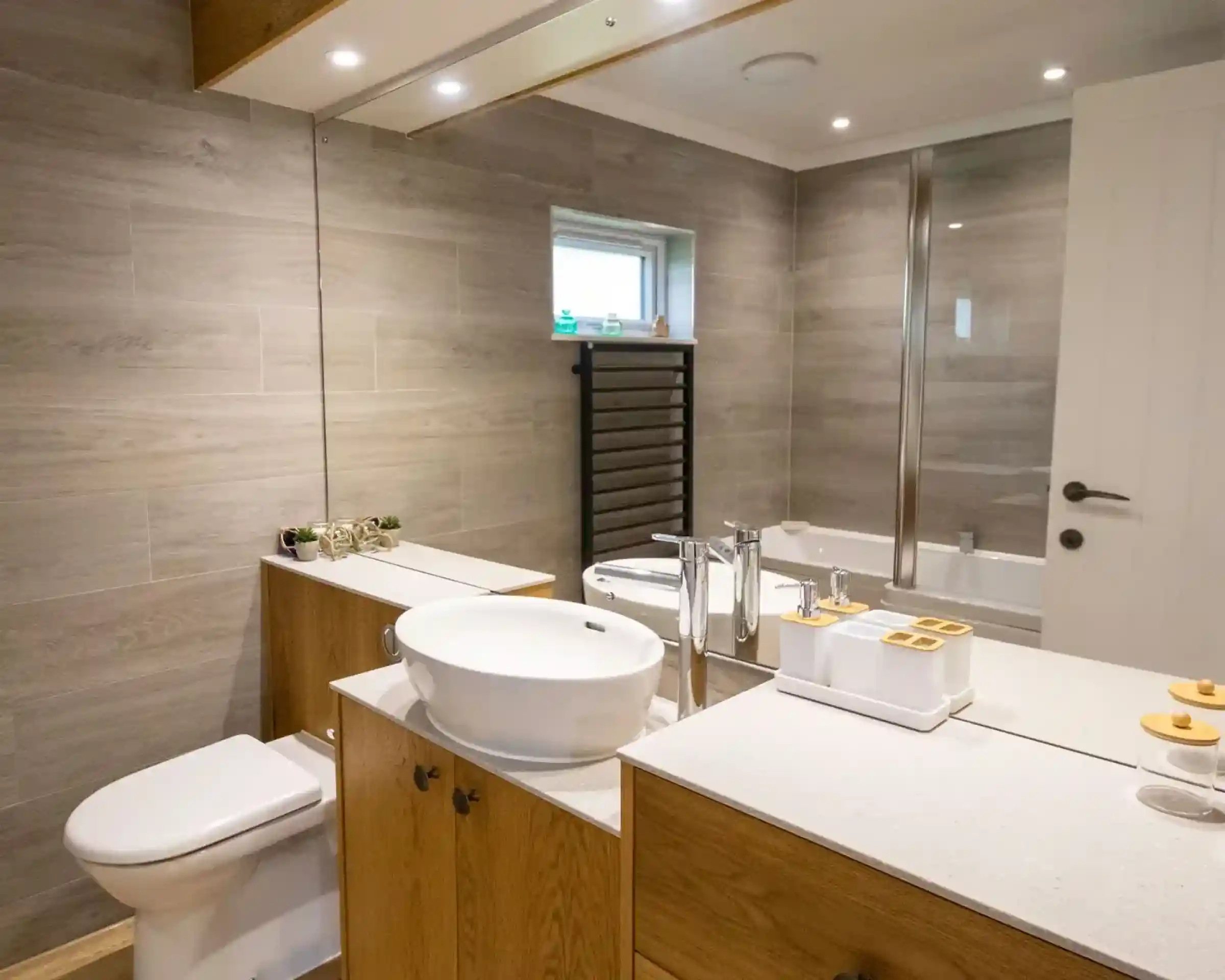 A modern bathroom features a wooden vanity with a round sink, a mirror above, and decorative items. There’s a toilet and a glass shower enclosure, along with gray tiled walls and a small window.
