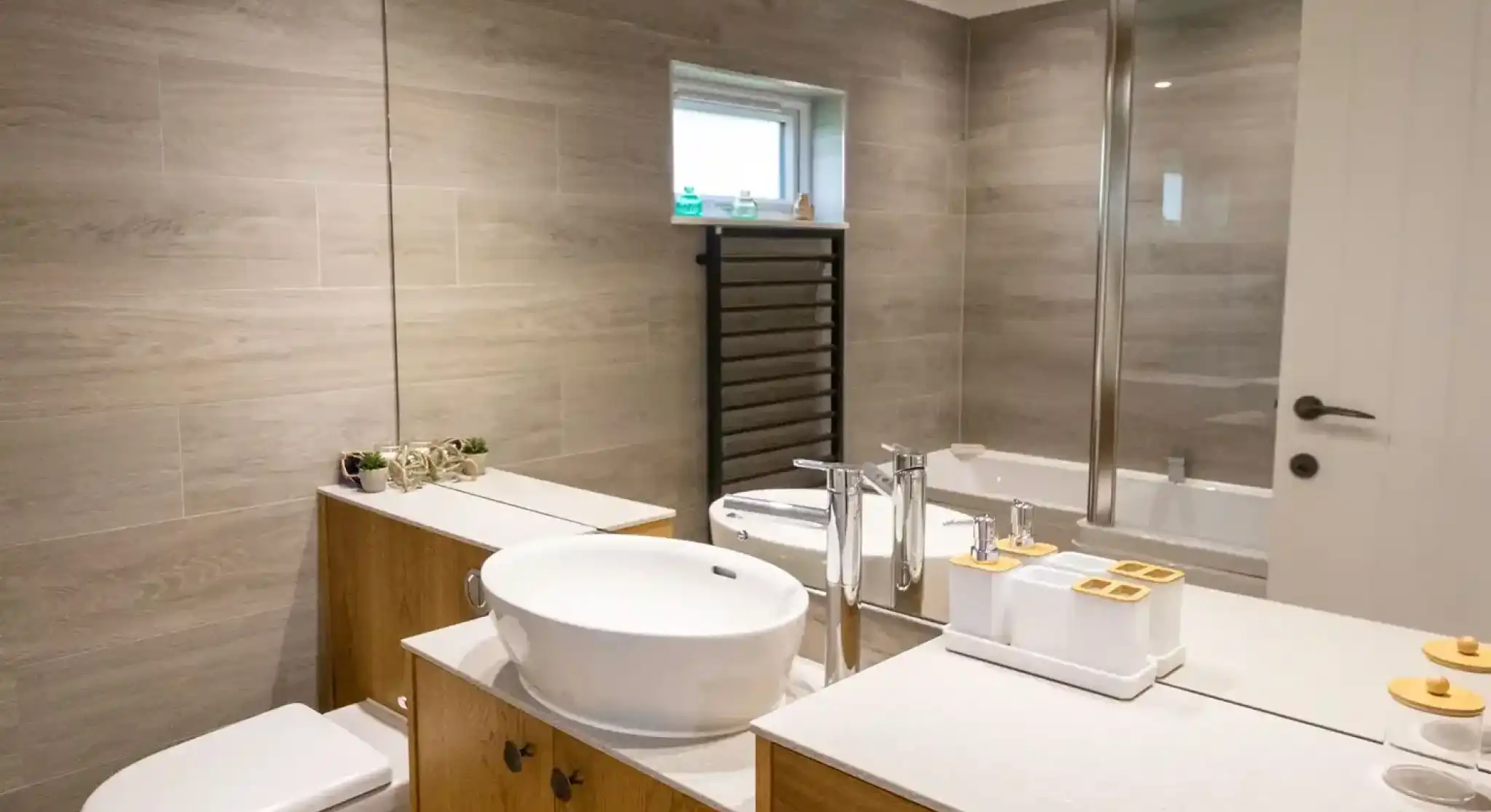 A modern bathroom features a wooden vanity with a round sink, a mirror above, and decorative items. There’s a toilet and a glass shower enclosure, along with gray tiled walls and a small window.