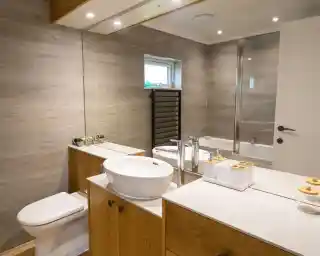 A modern bathroom features a wooden vanity with a round sink, a mirror above, and decorative items. There’s a toilet and a glass shower enclosure, along with gray tiled walls and a small window.