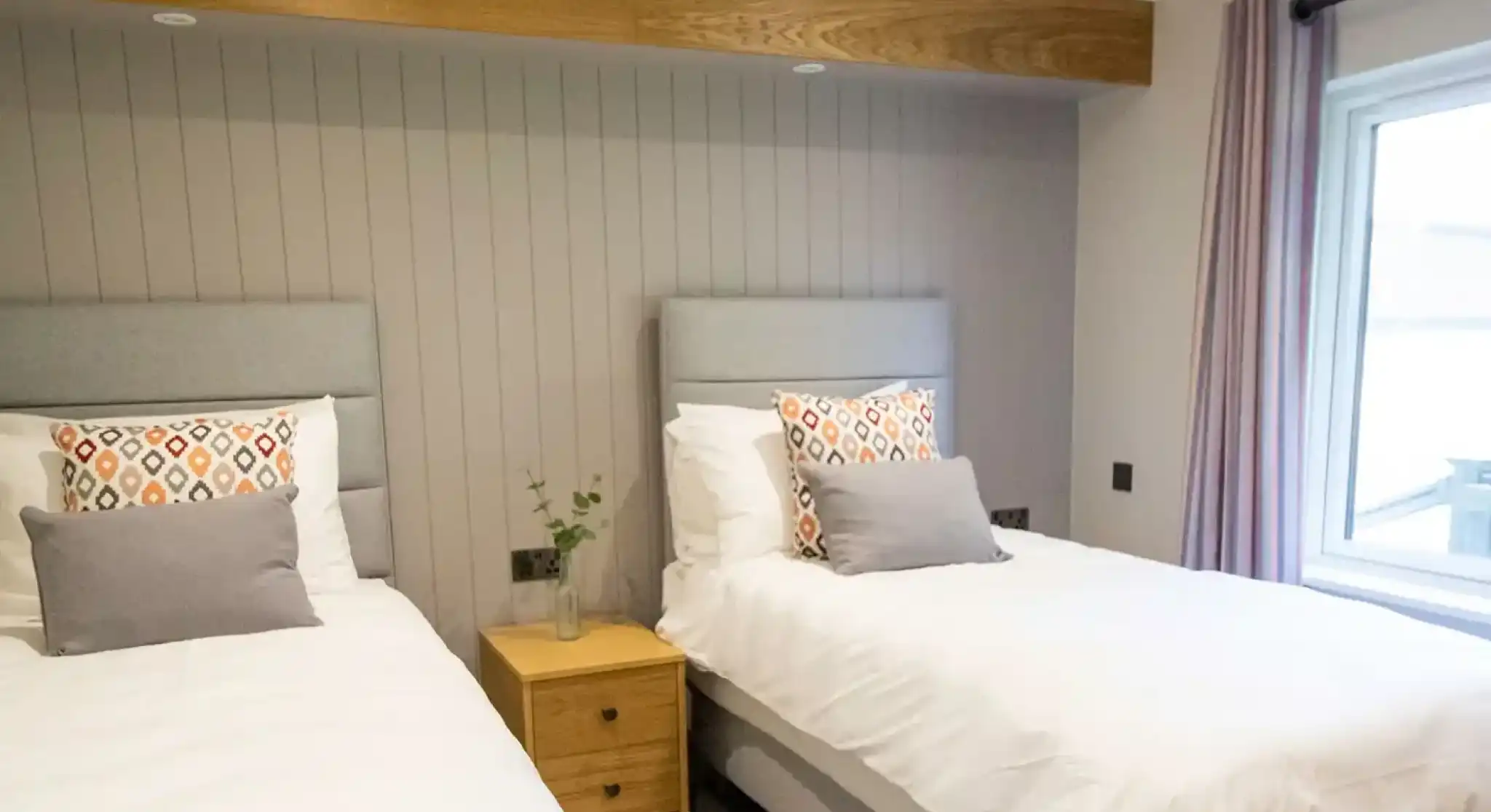 Two twin beds with grey headboards and patterned pillows in a room with wood accents and a large window.