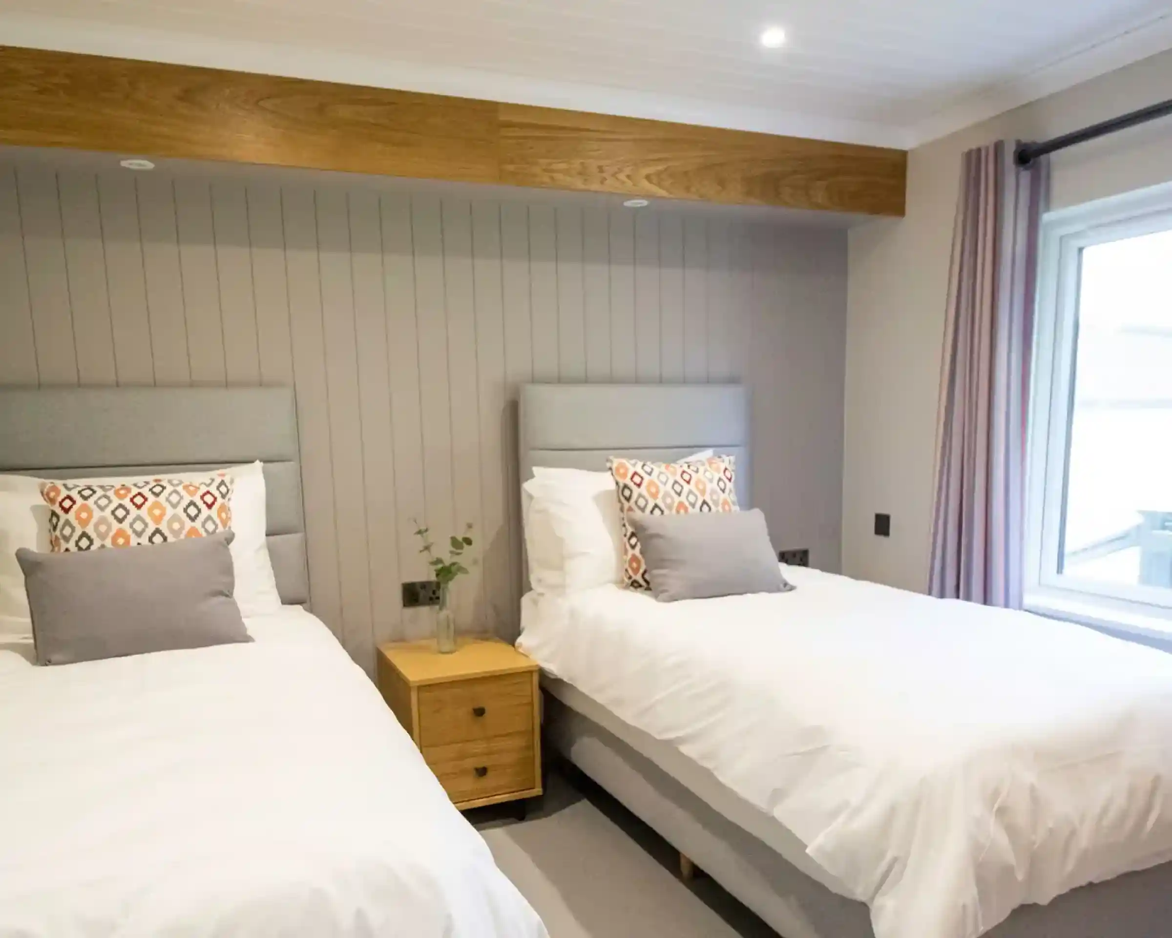 Two twin beds with grey headboards and patterned pillows in a room with wood accents and a large window.