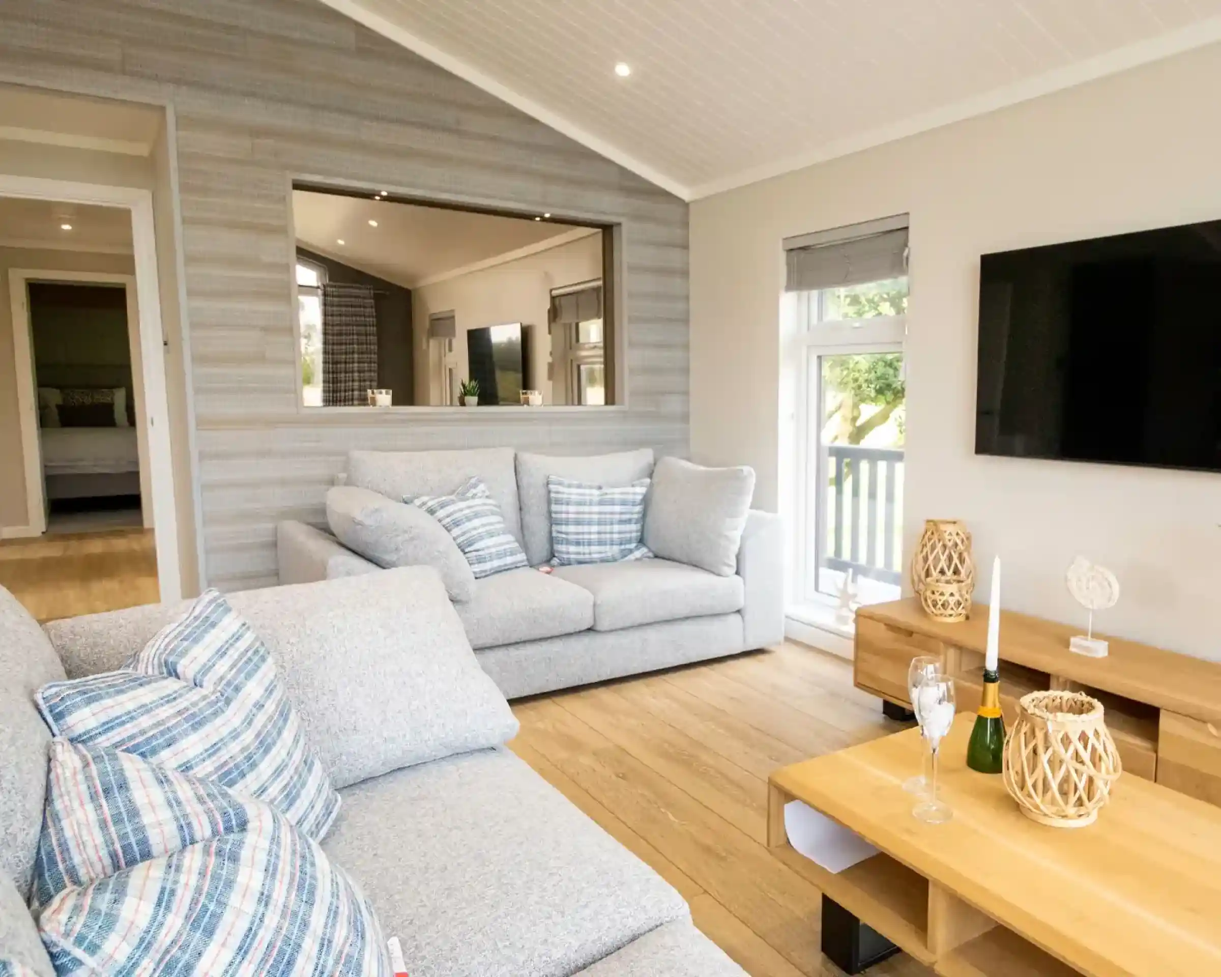 A bright, modern living room with a grey sectional sofa, a large television, and a wooden coffee table with champagne flutes.