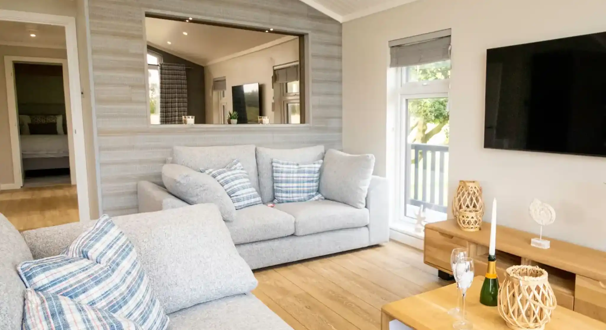 A bright, modern living room with a grey sectional sofa, a large television, and a wooden coffee table with champagne flutes.