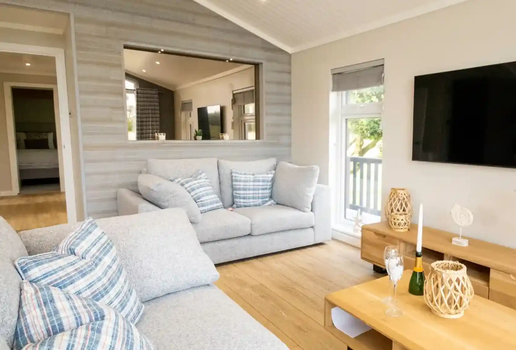 A bright, modern living room with a grey sectional sofa, a large television, and a wooden coffee table with champagne flutes.