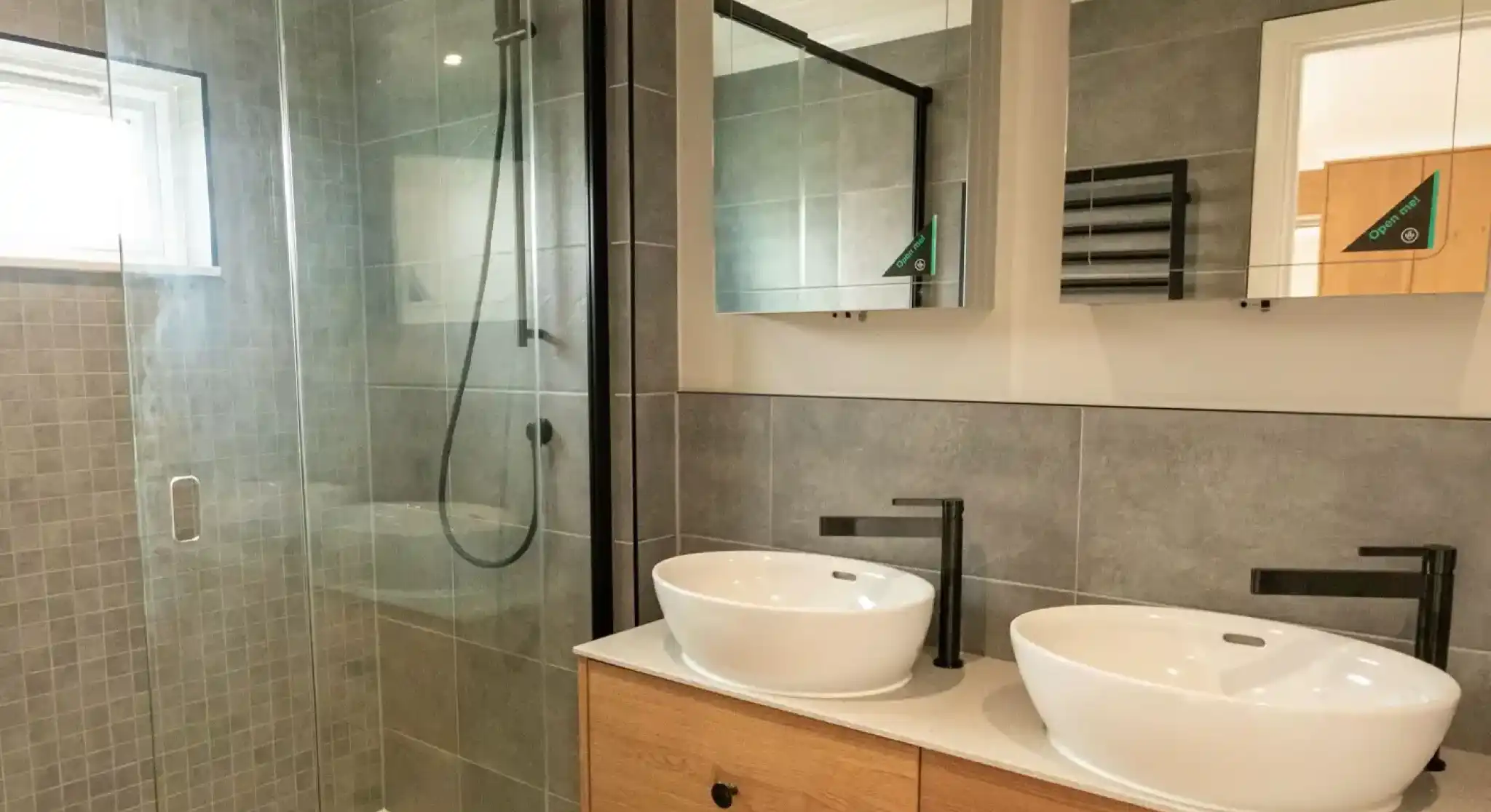 Modern bathroom with a glass-enclosed shower, two white vessel sinks, and wooden vanity cabinets.