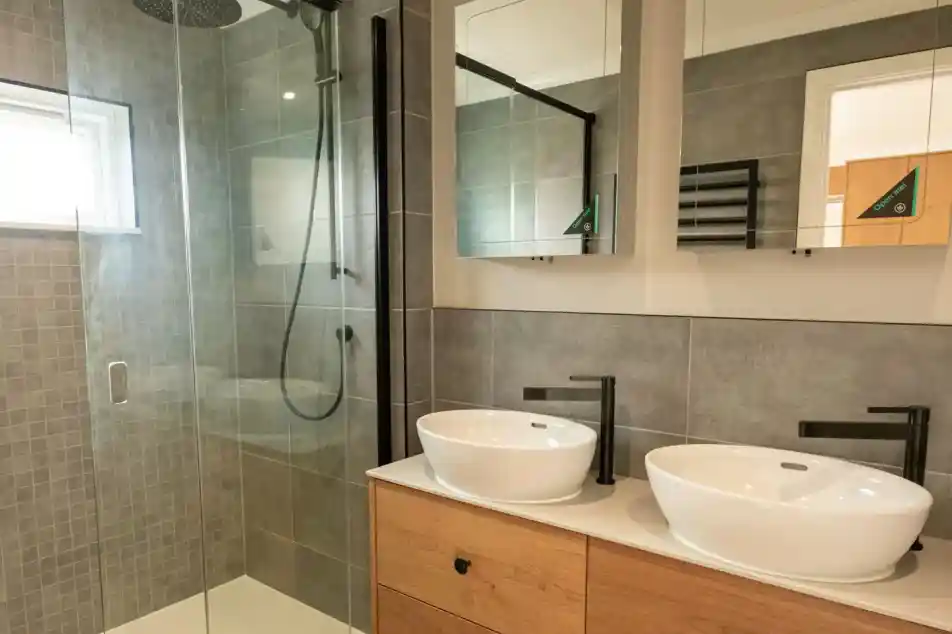 Modern bathroom with a glass-enclosed shower, two white vessel sinks, and wooden vanity cabinets.
