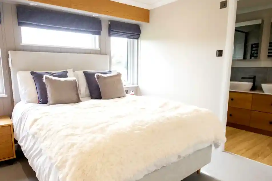 A large double bedroom featuring a plush white duvet, grey headboard, and a glimpse of a modern bathroom with double sinks.