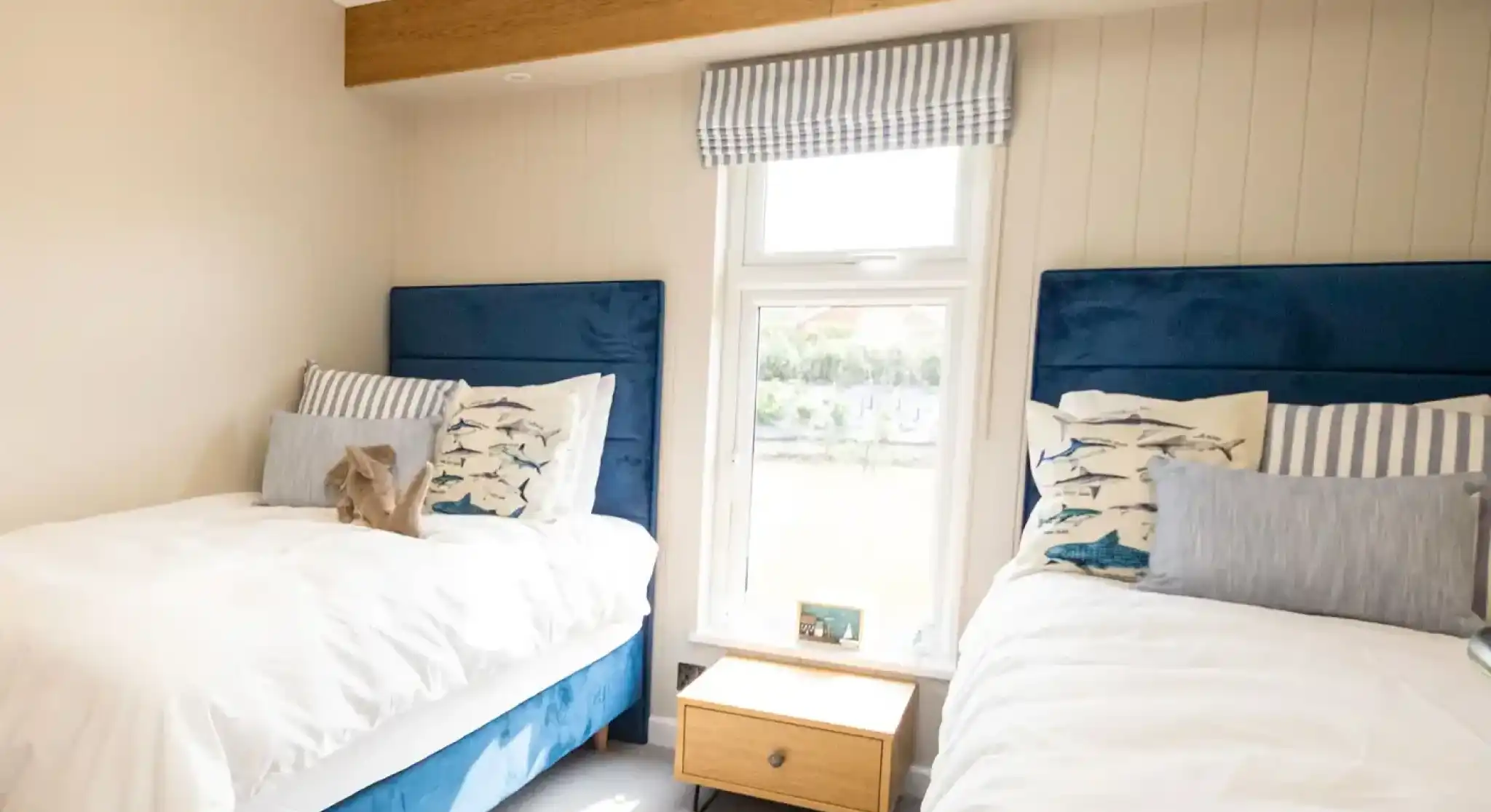 A cozy bedroom featuring two twin beds with blue upholstered headboards, dressed in white linens and decorative pillows. A small wooden nightstand sits between the beds, and a window offers a view, covered by striped curtains.