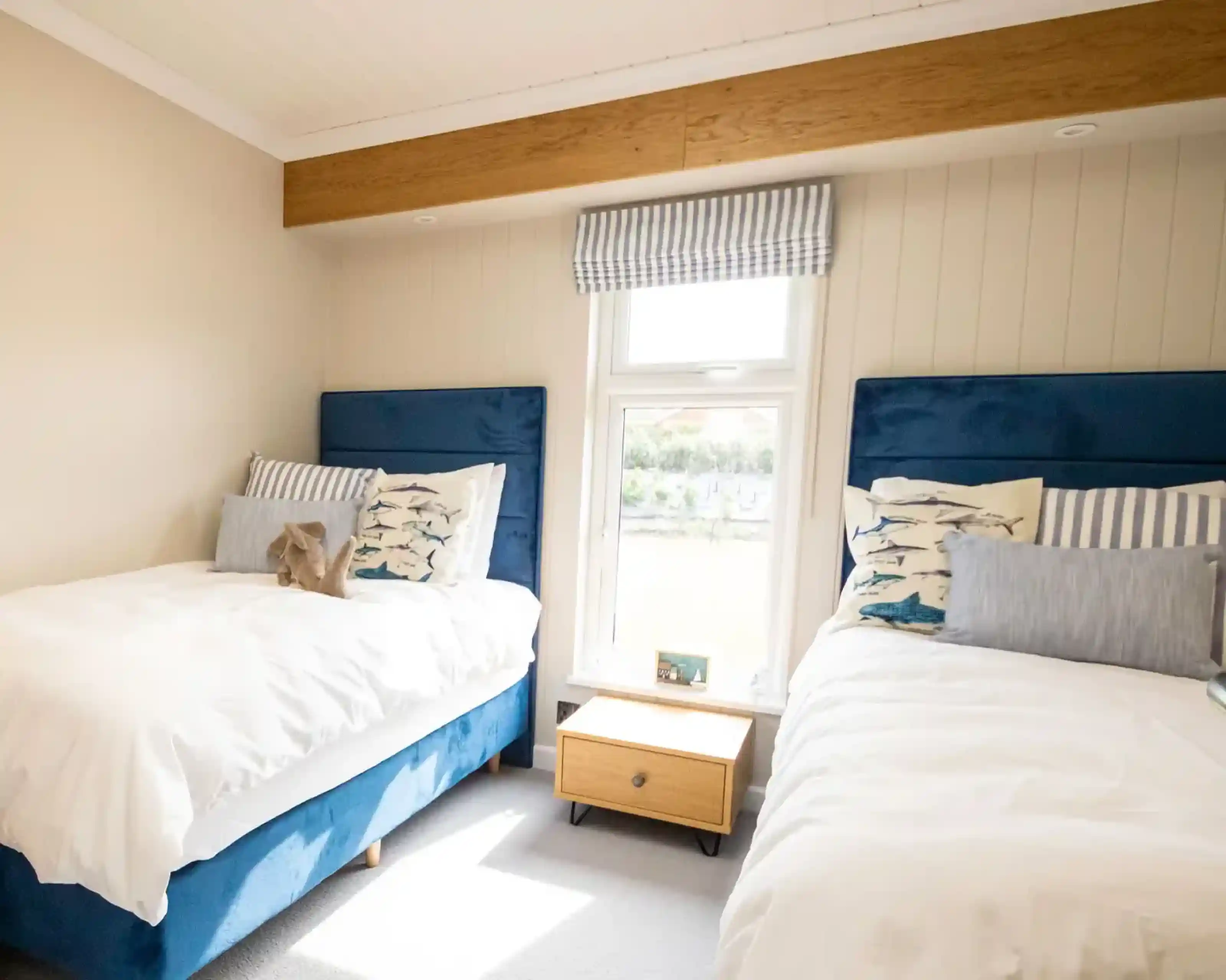 A cozy bedroom featuring two twin beds with blue upholstered headboards, dressed in white linens and decorative pillows. A small wooden nightstand sits between the beds, and a window offers a view, covered by striped curtains.