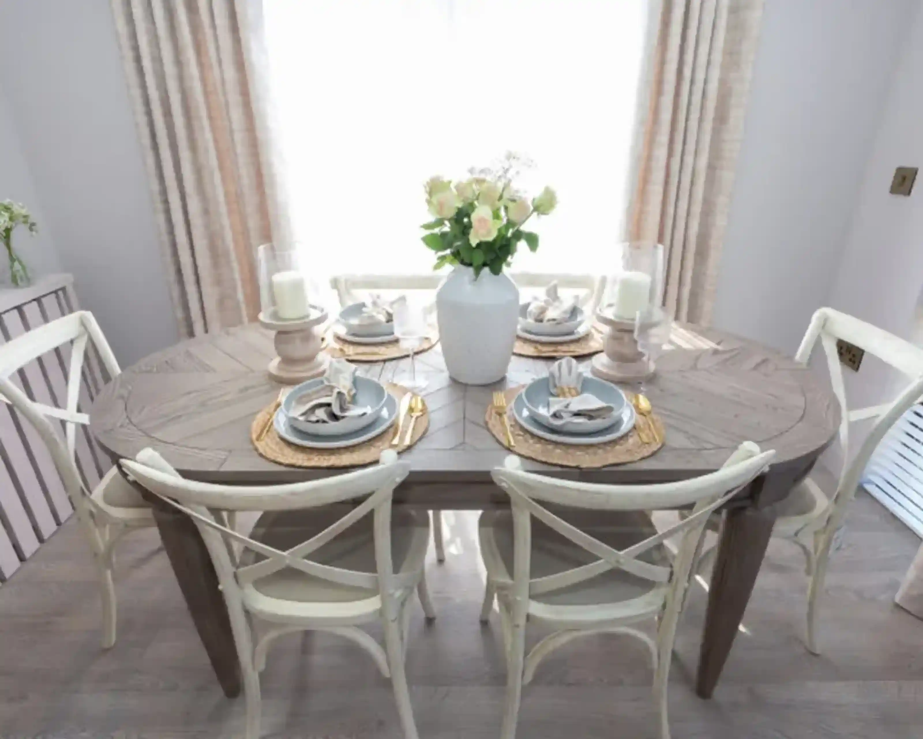 A dining table set for six with a vase of white roses and candlelight.