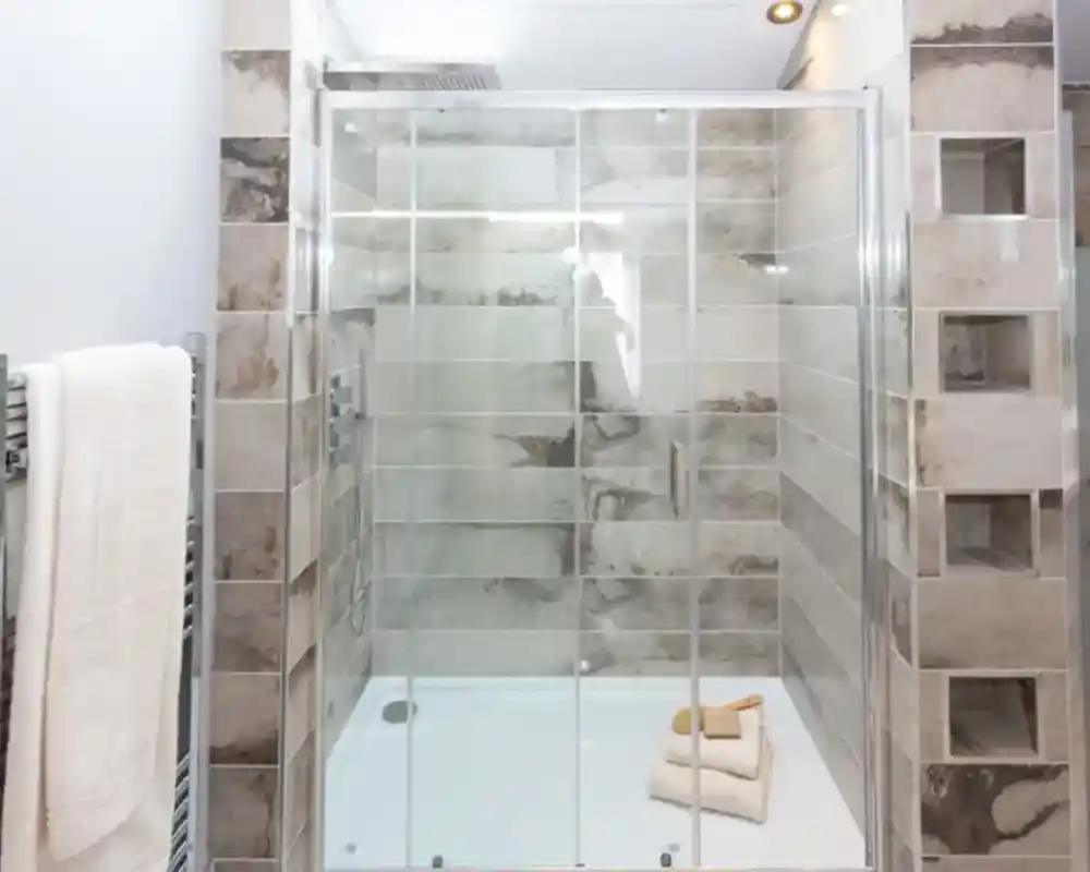 A modern shower enclosure with glass doors, tiled walls, a white shower base, and a towel rack with white towels.
