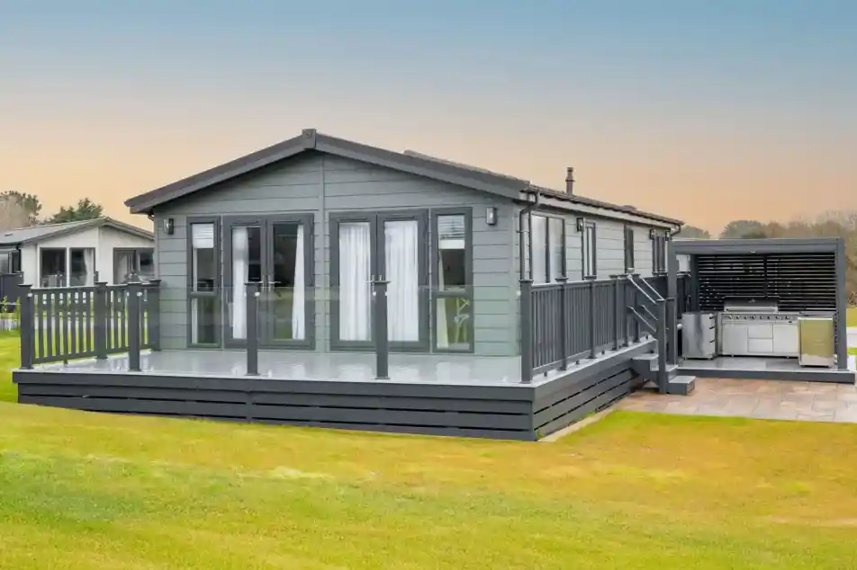 A modern grey lodge holiday home with a large deck and outdoor kitchen area sits on a grassy lawn under a pale blue sky.