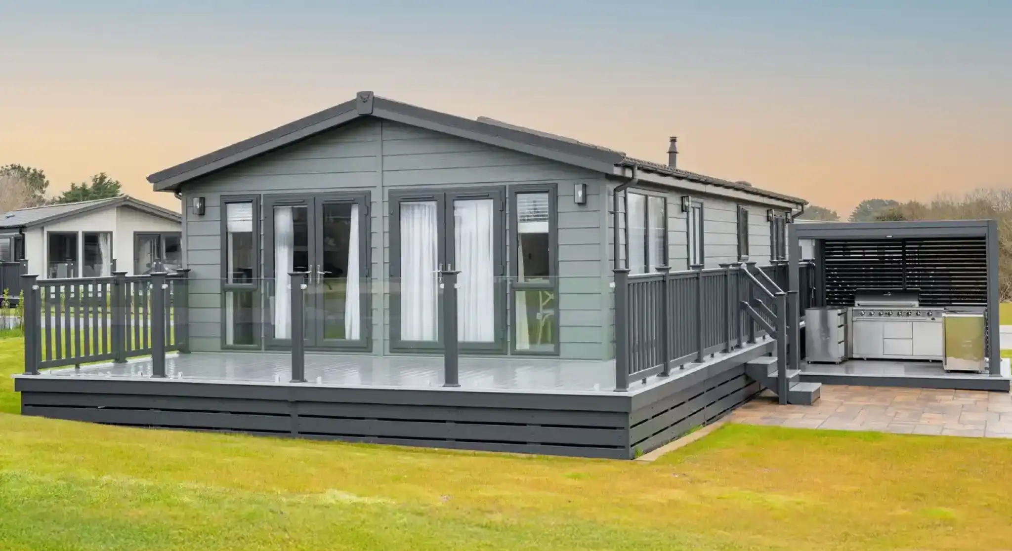 A modern grey lodge holiday home with a large deck and outdoor kitchen area sits on a grassy lawn under a pale blue sky.