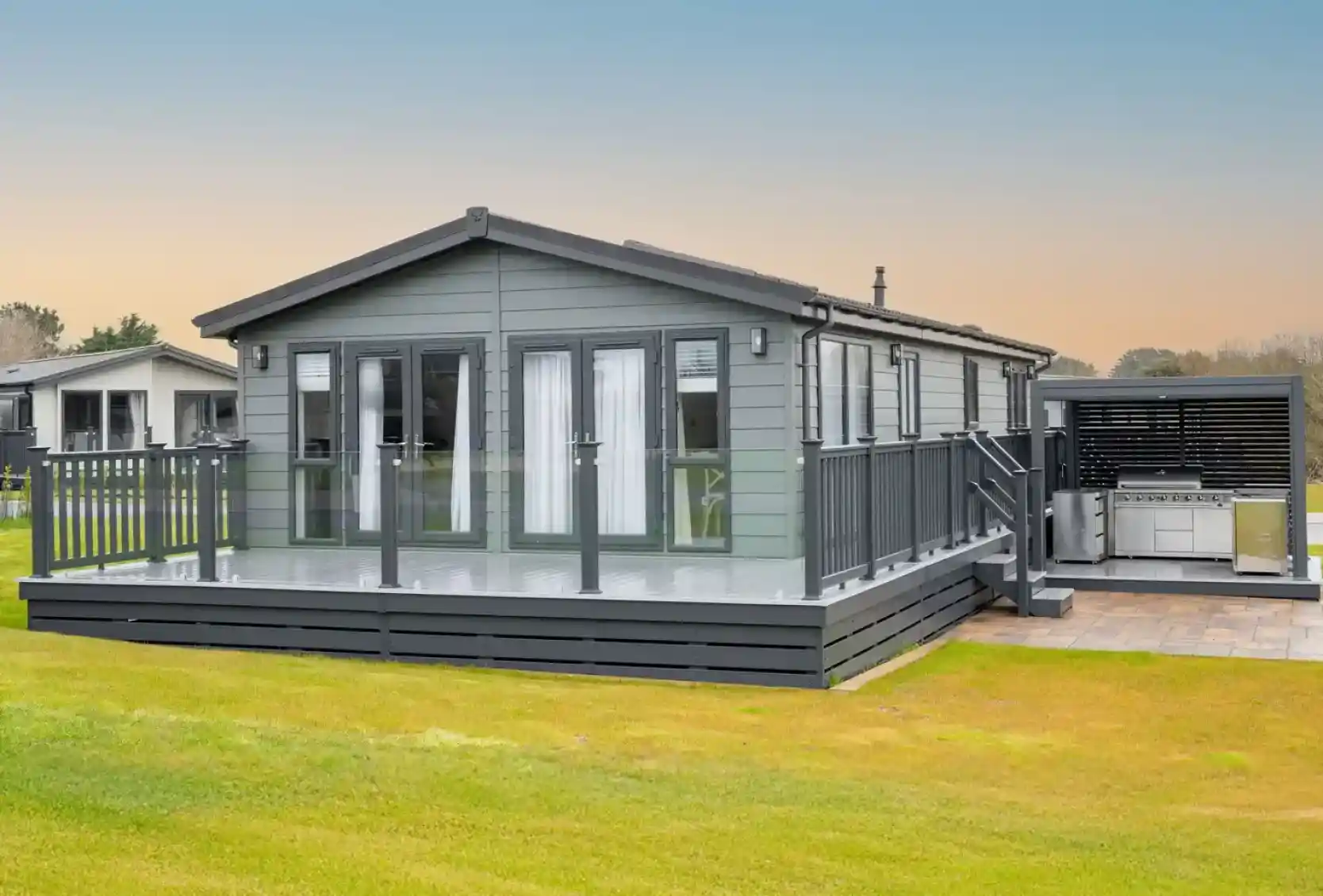 A modern grey lodge holiday home with a large deck and outdoor kitchen area sits on a grassy lawn under a pale blue sky.