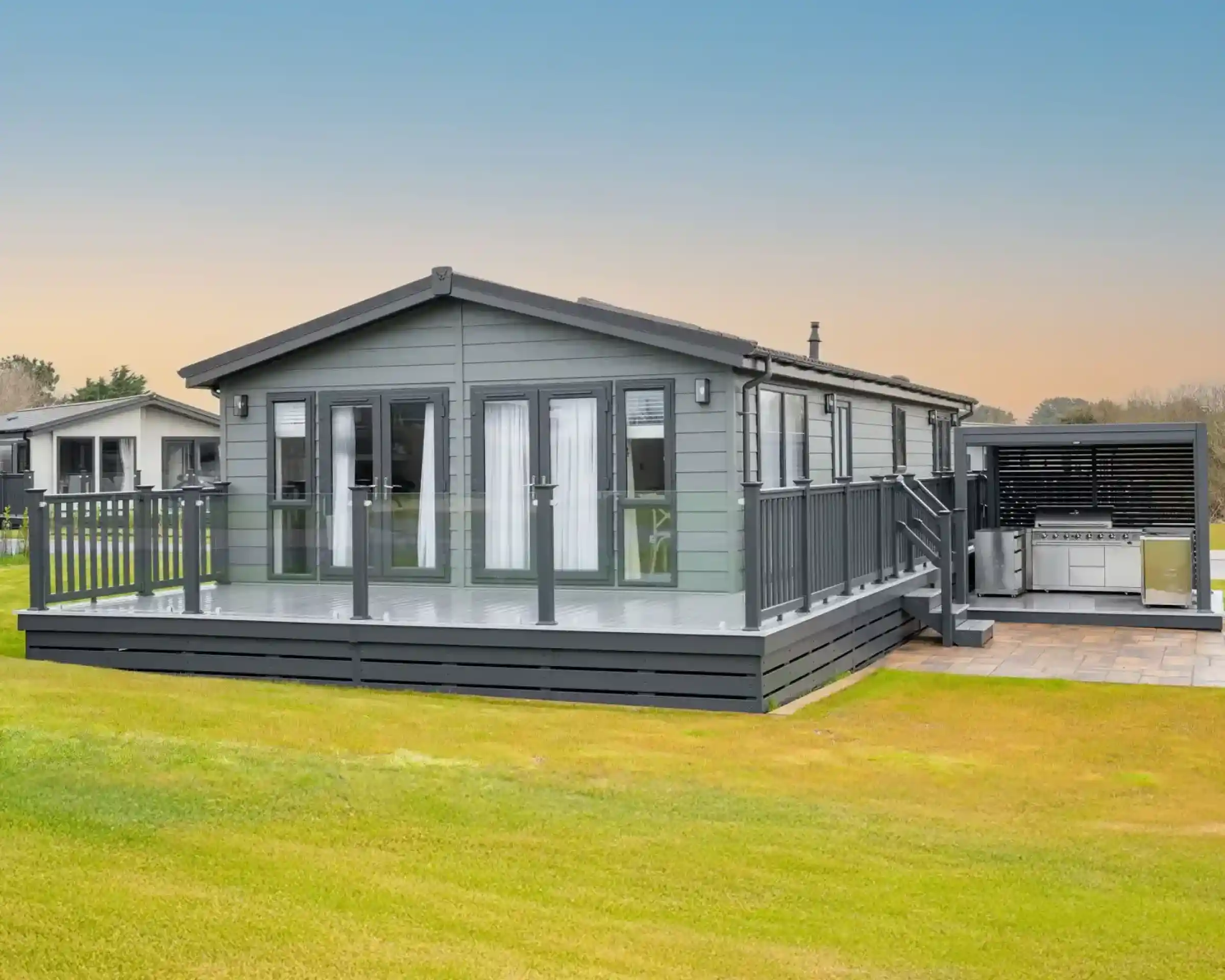 A modern grey lodge holiday home with a large deck and outdoor kitchen area sits on a grassy lawn under a pale blue sky.