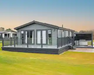 A modern grey lodge holiday home with a large deck and outdoor kitchen area sits on a grassy lawn under a pale blue sky.