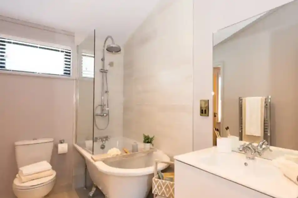 A modern bathroom features a freestanding clawfoot tub with a glass shower screen, a toilet with stacked towels, and a white vanity with a sink and mirror.