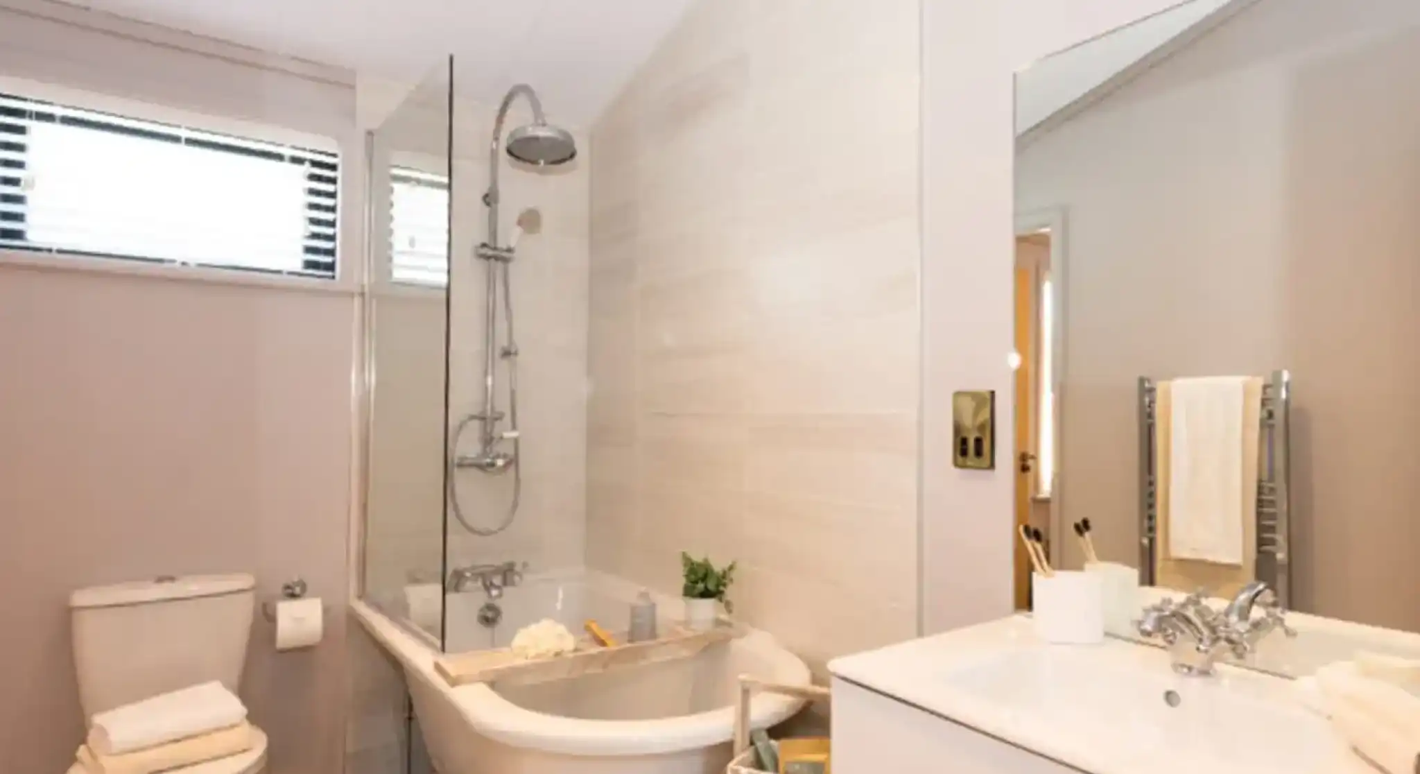 A modern bathroom features a freestanding clawfoot tub with a glass shower screen, a toilet with stacked towels, and a white vanity with a sink and mirror.
