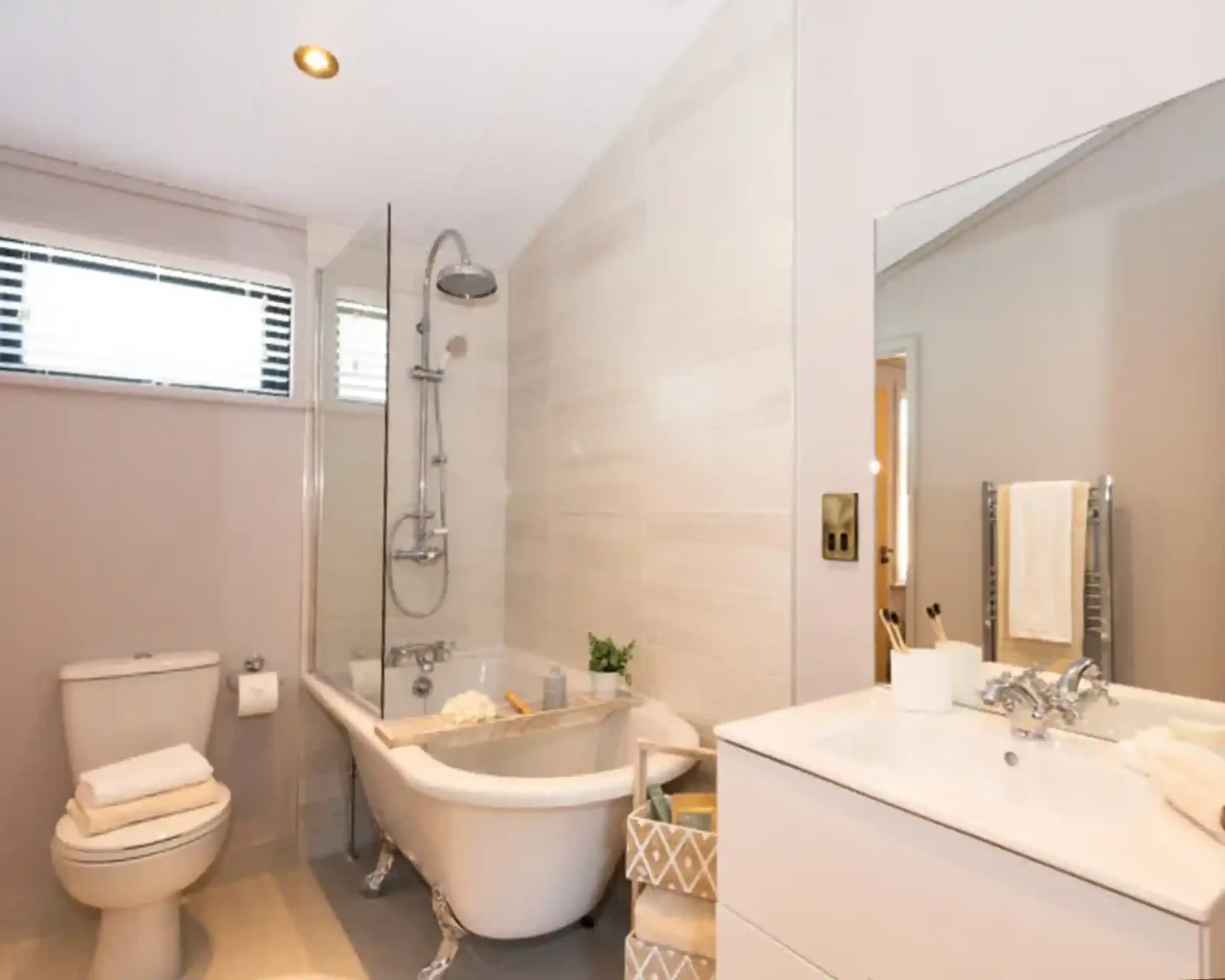 A modern bathroom features a freestanding clawfoot tub with a glass shower screen, a toilet with stacked towels, and a white vanity with a sink and mirror.