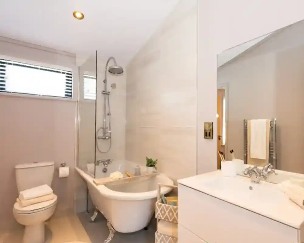 A modern bathroom features a freestanding clawfoot tub with a glass shower screen, a toilet with stacked towels, and a white vanity with a sink and mirror.