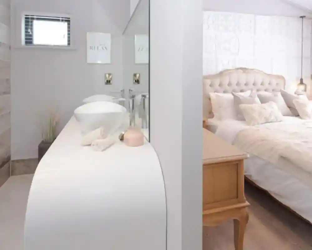Interior view of a modern bathroom vanity area with two sinks and a glimpse of a tufted headboard bedroom.
