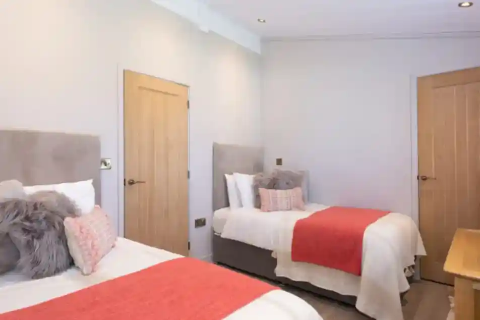 Two beds with coral throws and plush pillows in a modern bedroom with light grey walls and natural wood doors.