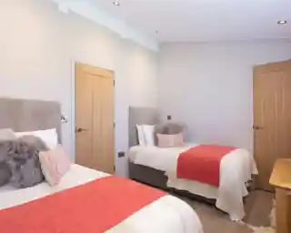 Two beds with coral throws and plush pillows in a modern bedroom with light grey walls and natural wood doors.