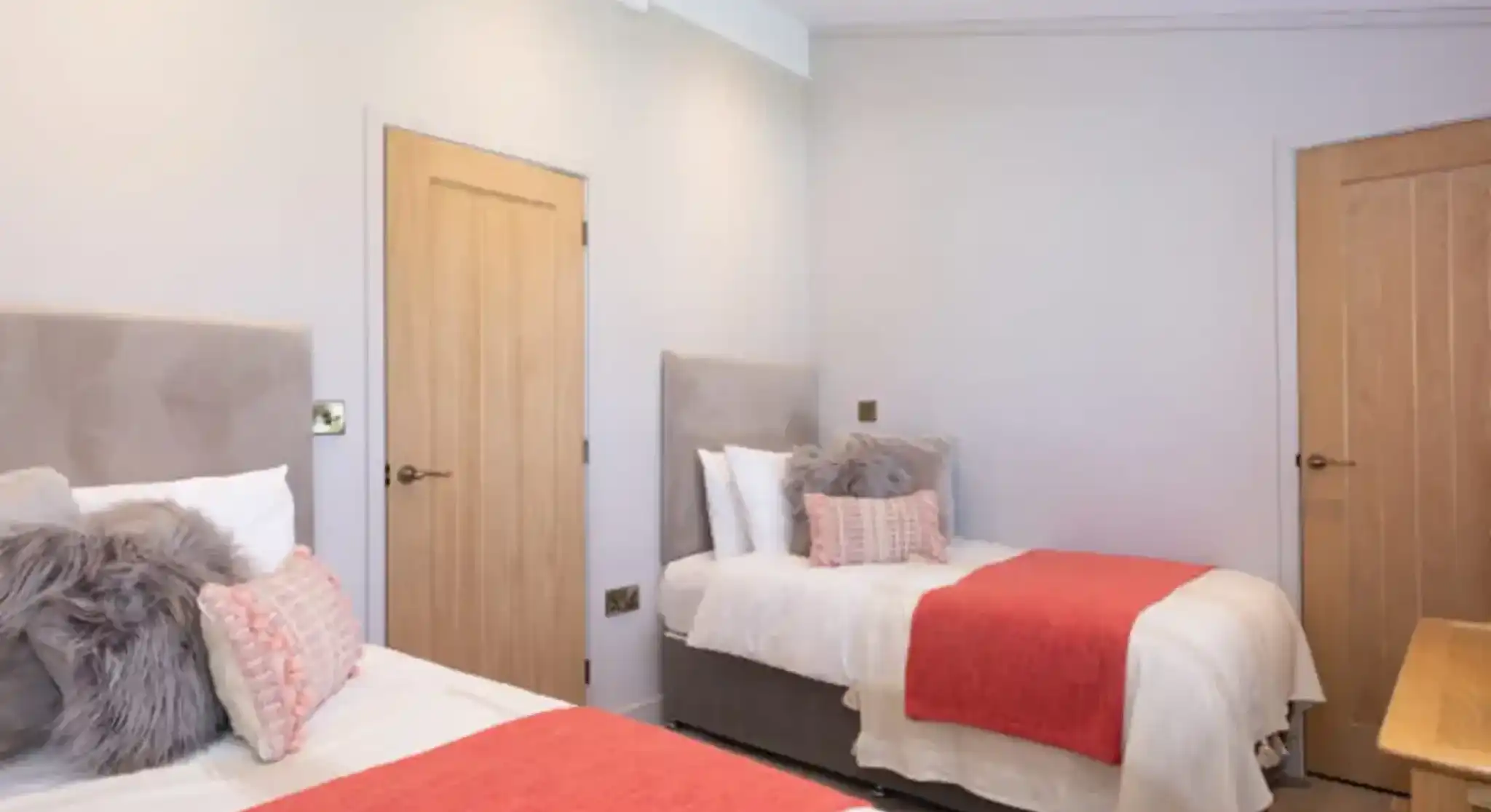 Two beds with coral throws and plush pillows in a modern bedroom with light grey walls and natural wood doors.