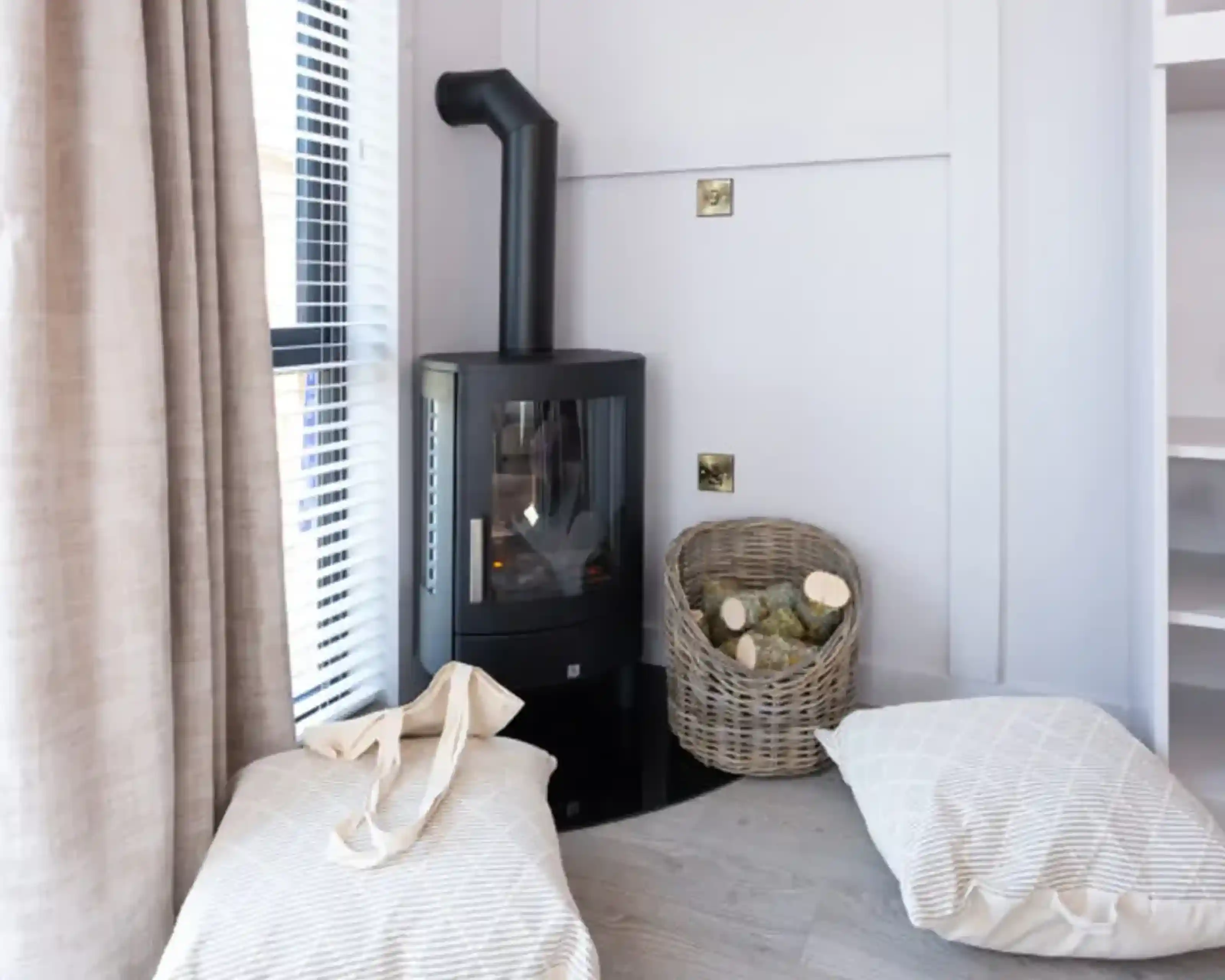 A modern wood stove is situated in a cozy corner next to a basket of firewood. Two soft, striped cushions are placed on the floor near the stove, and sunlight filters through a window with blinds.