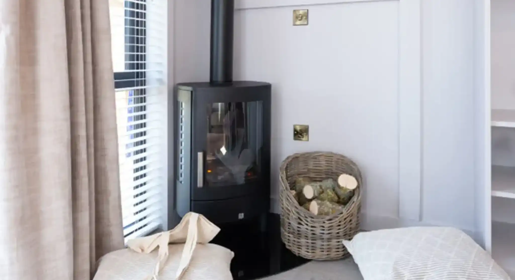 A modern wood stove is situated in a cozy corner next to a basket of firewood. Two soft, striped cushions are placed on the floor near the stove, and sunlight filters through a window with blinds.