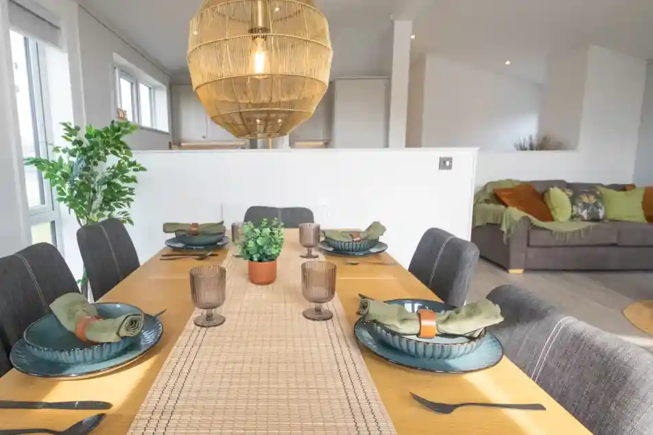A modern dining area with a wooden table set for a meal, featuring dark grey chairs, woven placemats, blue dishes, and a large wicker pendant light overhead.