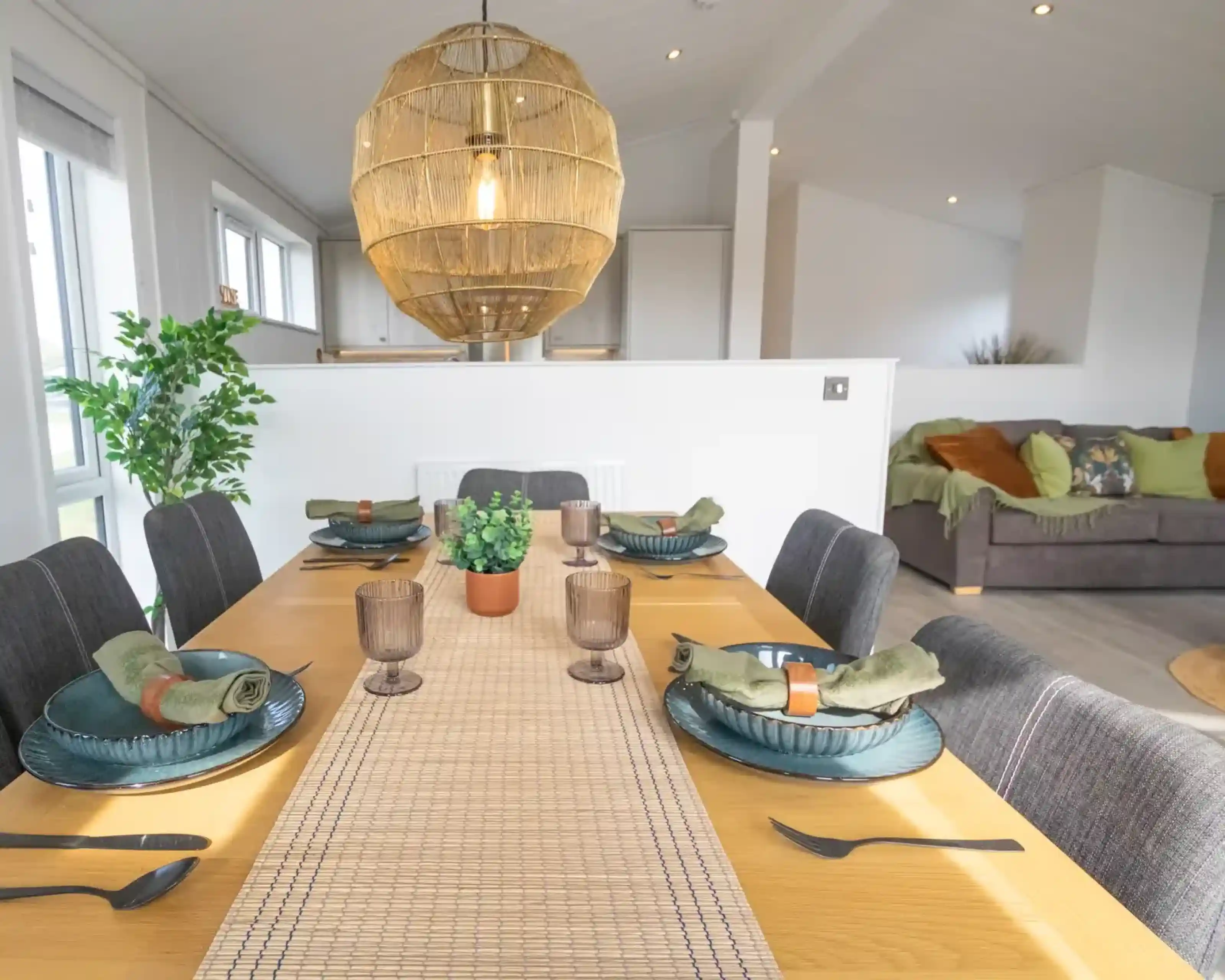 A modern dining area with a wooden table set for a meal, featuring dark grey chairs, woven placemats, blue dishes, and a large wicker pendant light overhead.