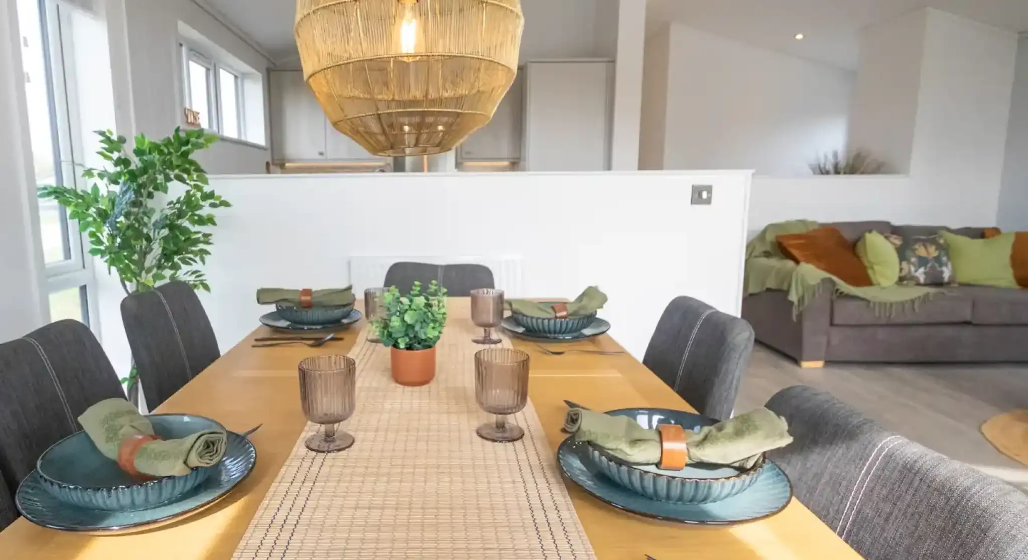 A modern dining area with a wooden table set for a meal, featuring dark grey chairs, woven placemats, blue dishes, and a large wicker pendant light overhead.