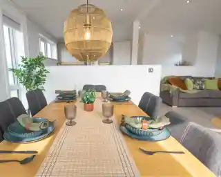 A modern dining area with a wooden table set for a meal, featuring dark grey chairs, woven placemats, blue dishes, and a large wicker pendant light overhead.