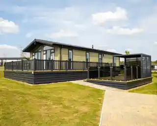 Modern light-green mobile home with a large wooden deck and black railings, adjacent to a covered outdoor seating area.