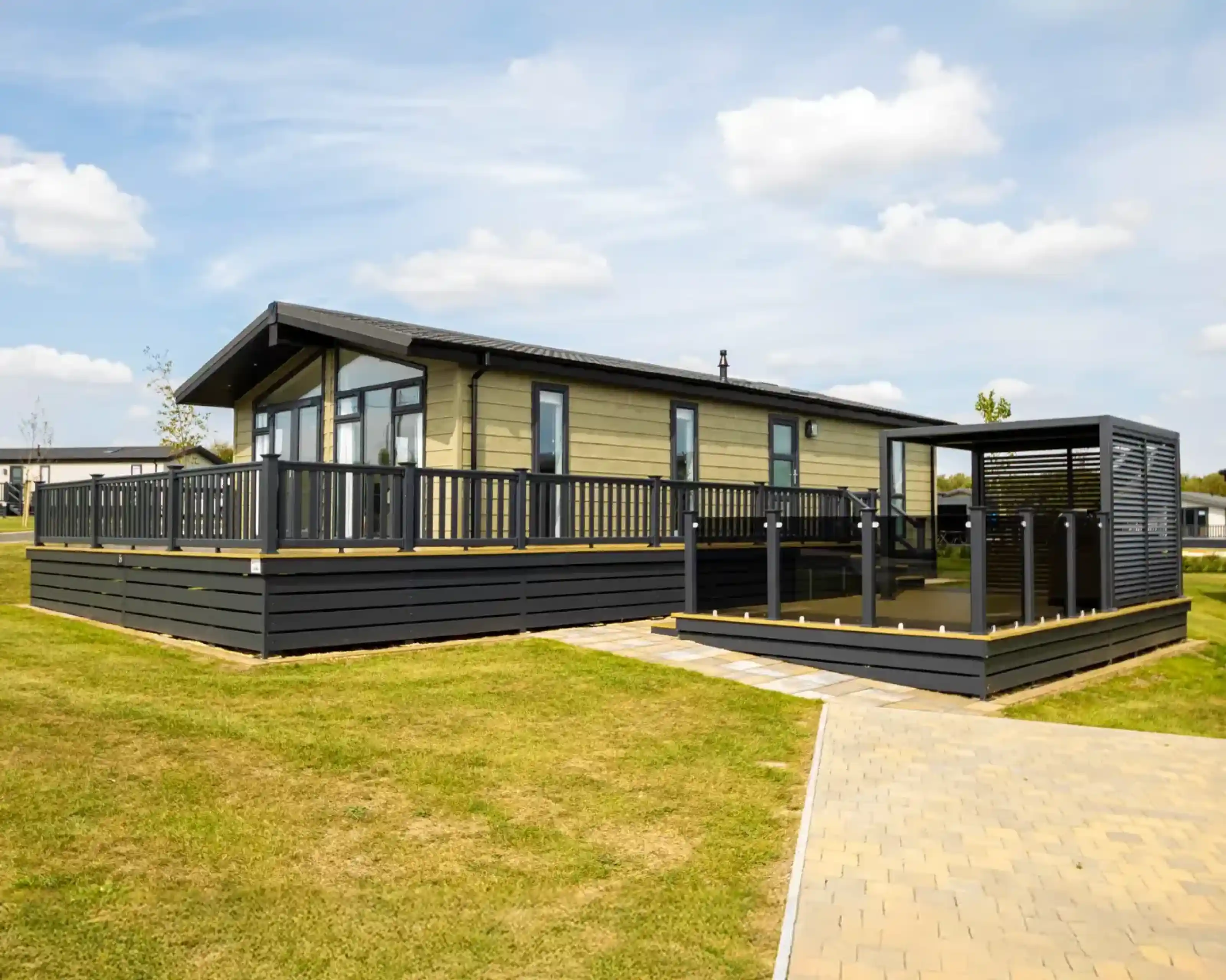Modern light-green mobile home with a large wooden deck and black railings, adjacent to a covered outdoor seating area.