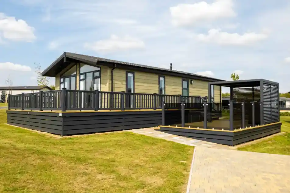 Modern light-green mobile home with a large wooden deck and black railings, adjacent to a covered outdoor seating area.